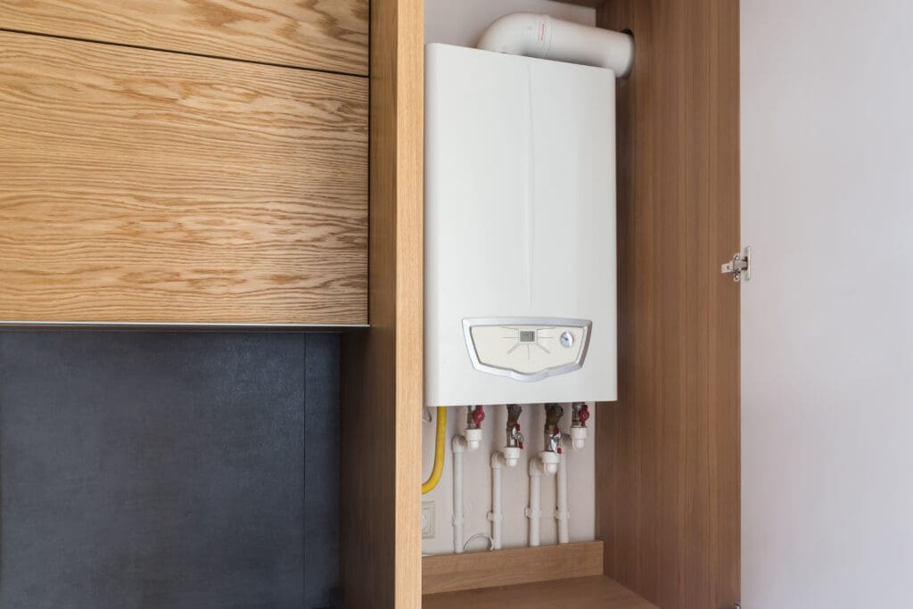 A white wall-mounted boiler with visible pipes is installed inside a wooden cabinet, with the cabinet door partially open. The setup is next to wooden drawers and a dark-colored panel.