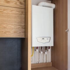 A white wall-mounted boiler with visible pipes is installed inside a wooden cabinet, with the cabinet door partially open. The setup is next to wooden drawers and a dark-colored panel.