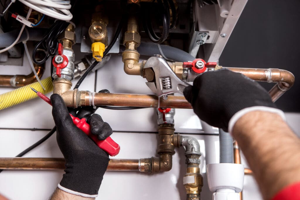 A person wearing black gloves uses a wrench and screwdriver to adjust copper pipes and valves on a plumbing system with various fittings and connections.