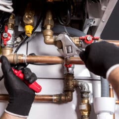 A person wearing black gloves uses a wrench and screwdriver to adjust copper pipes and valves on a plumbing system with various fittings and connections.