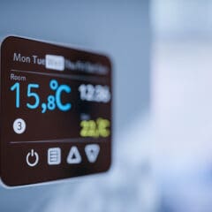 A close-up of a digital thermostat mounted on a wall displays the room temperature as 15.8°C, with other controls and icons visible on its screen. The background is blurred.