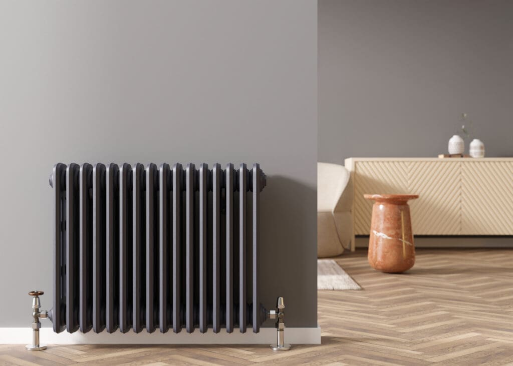 A black metal radiator is mounted on a gray wall in a modern room with herringbone wood flooring. A beige cabinet, orange marble side table, and light sofa are visible in the bright background.