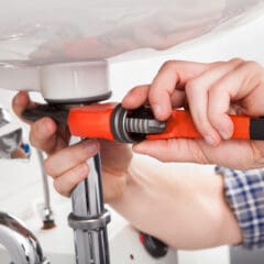 A person uses a red pipe wrench to tighten or loosen the pipe connections under a white bathroom sink, performing plumbing repairs.