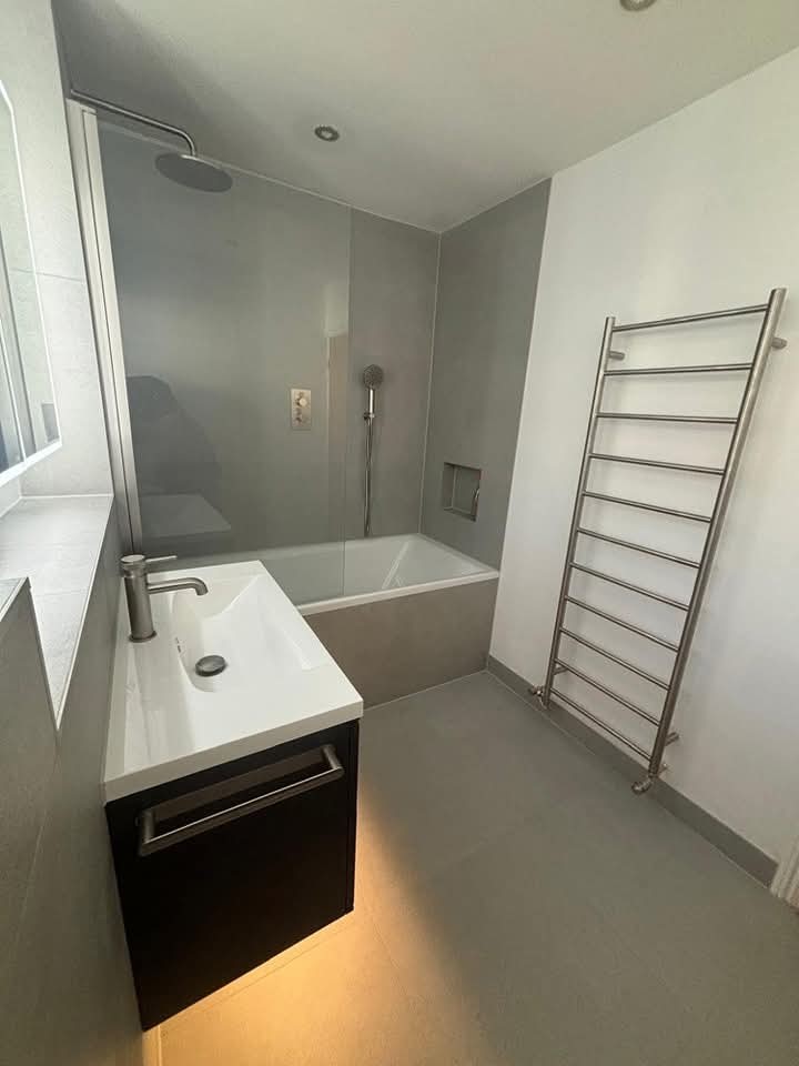 Modern bathroom with a wall-mounted sink, large mirror, bathtub with overhead shower, gray wall tiles, towel radiator, and minimalistic design. Natural light enters from a window on the left.
