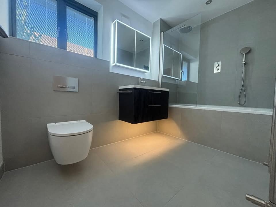 Modern bathroom with grey tiles, wall-mounted toilet, floating sink cabinet with underlighting, illuminated mirror, and a glass-enclosed shower and bathtub under a window.