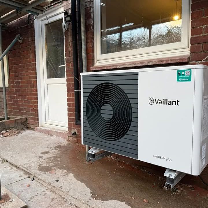A Vaillant aroTHERM plus heat pump is installed outside a brick building, near a white door. The unit is elevated on metal legs and sits on a concrete surface. Some outdoor wires and pipes are visible.