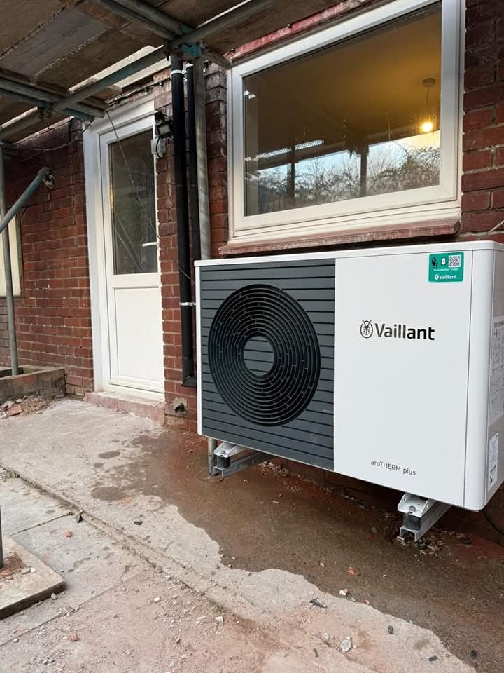 A Vaillant aroTHERM plus heat pump unit is installed outside a brick building, near a white-framed door and window, under a partially covered area with visible construction materials.