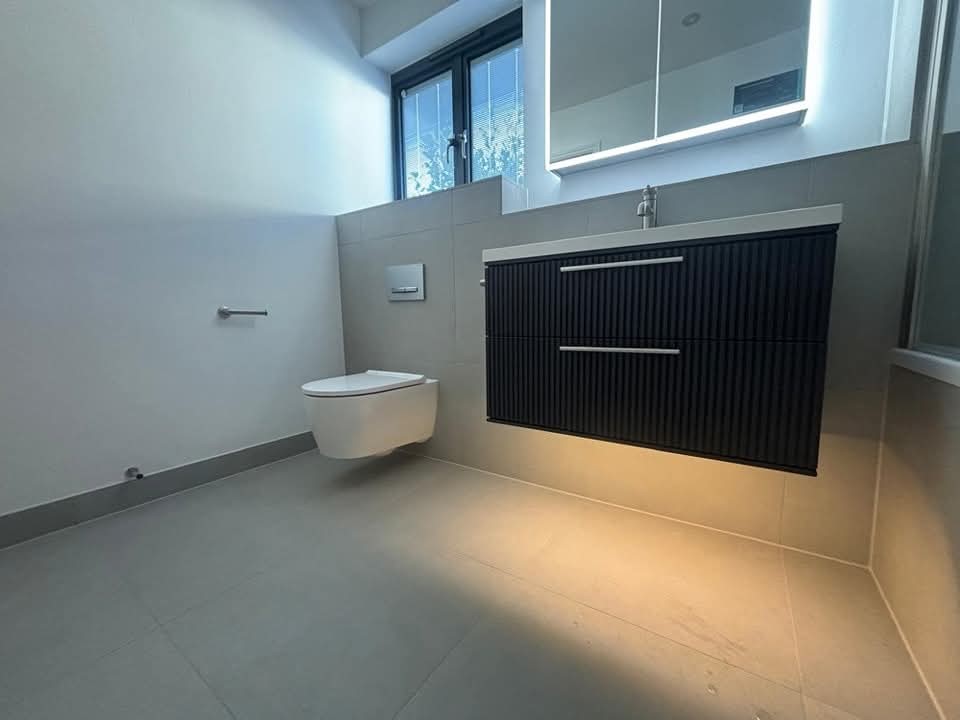 Modern bathroom with a wall-mounted toilet, floating vanity with two drawers, large mirror above the sink, and natural light coming from a window. Light shines under the vanity, illuminating the floor.