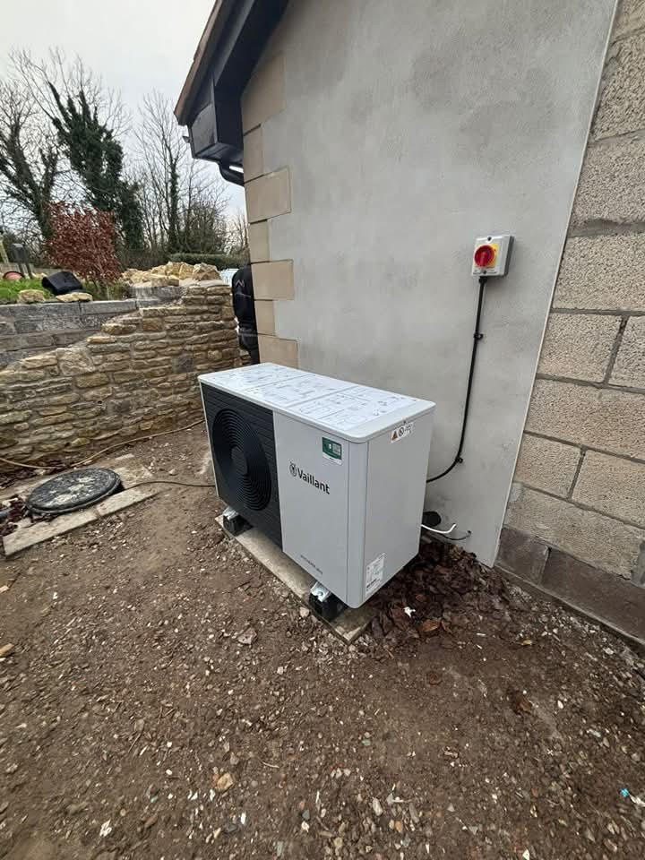 A Vaillant heat pump is installed outside a building on a concrete slab, with an electric switch mounted on the wall nearby and trees and a stone wall in the background.