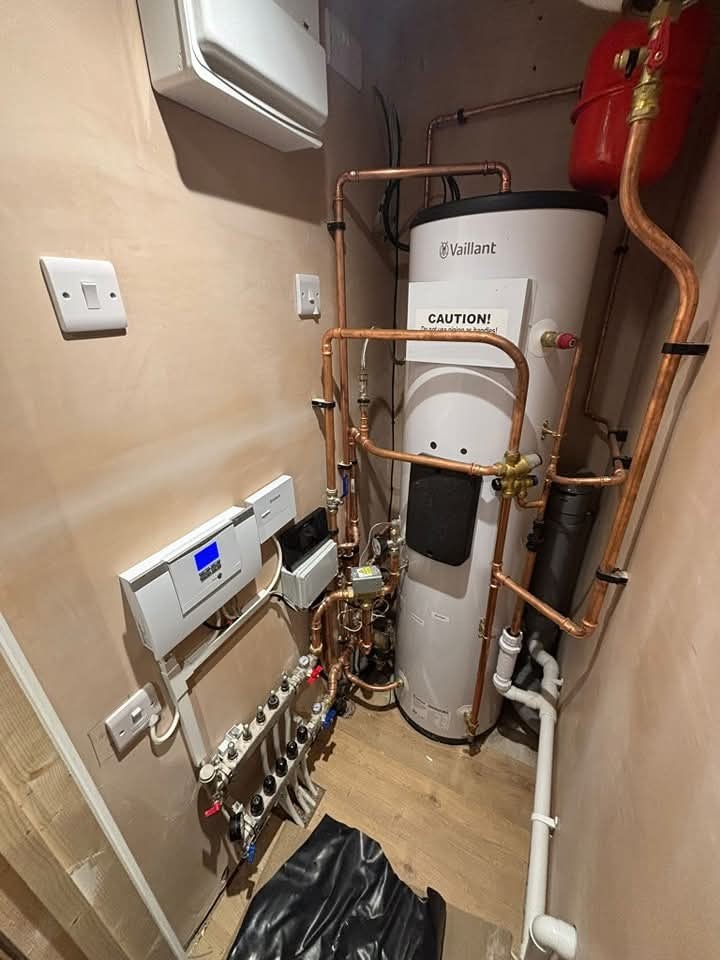 A compact utility room with a white Vaillant hot water cylinder, exposed copper pipes, control panels, and a red expansion vessel on the wall. The setup is tidy and modern, with a wooden floor and beige walls.