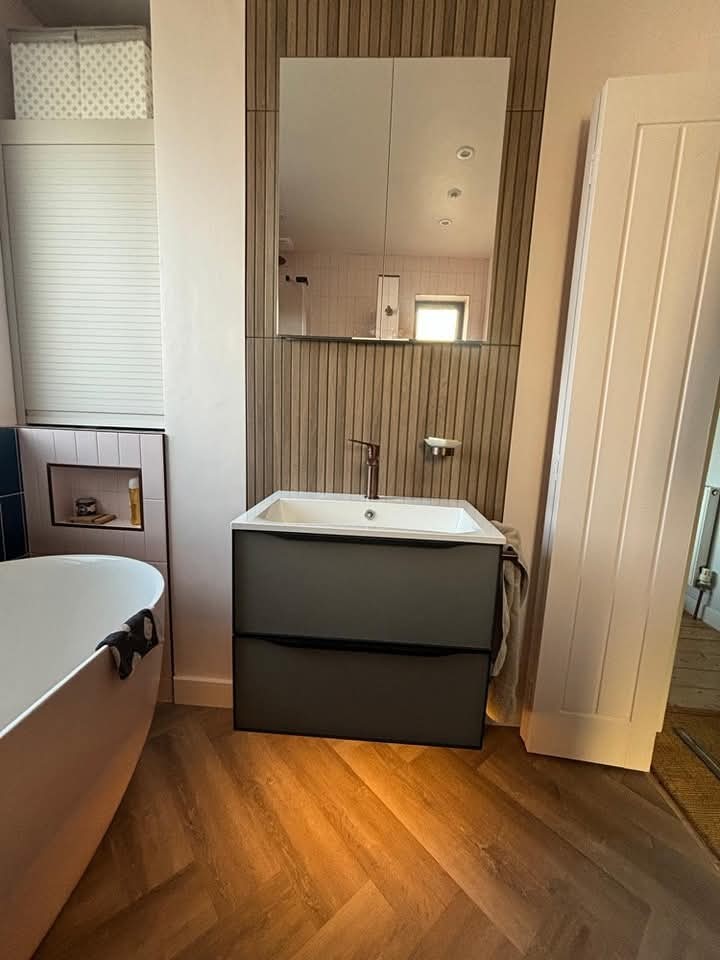 Modern bathroom with a freestanding white bathtub, a gray vanity with a white sink, large mirror above, and wood-patterned floor. There's a white door open to the right and small shelves on the left.