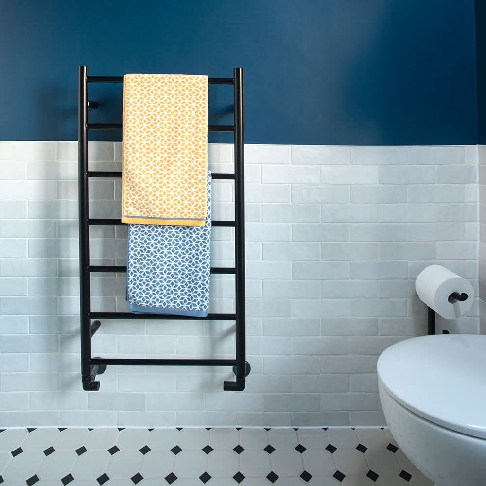 A black towel rack holds a yellow towel and a blue towel against a white tiled wall with a blue upper section. A white toilet and a toilet paper roll are visible on the right. The floor has black and white tiles.