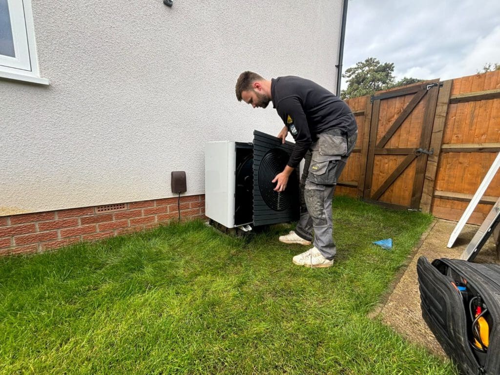 A man in work clothes installs or repairs an outdoor appliance next to a house, standing on grass near a wooden gate and a tool bag.