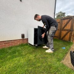 A man in work clothes installs or repairs an outdoor appliance next to a house, standing on grass near a wooden gate and a tool bag.