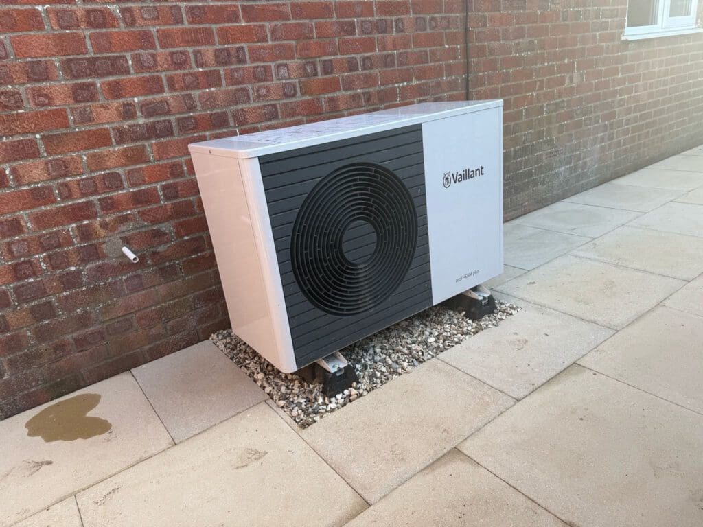 A Vaillant heat pump unit is installed outdoors on a stone bed against a brick wall, surrounded by beige paving slabs.