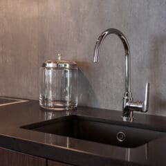 A modern kitchen sink with a tall, curved chrome faucet set in a dark countertop. There is a clear glass jar with a metal lid next to the sink, and the backsplash is a smooth, gray surface.