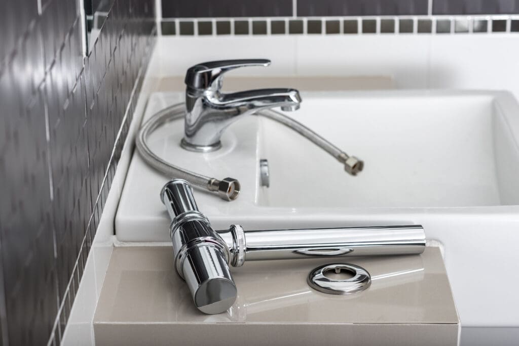 A chrome bathroom faucet and flexible hoses are placed on a counter next to a white sink, with shiny black and white tiles on the wall in the background.