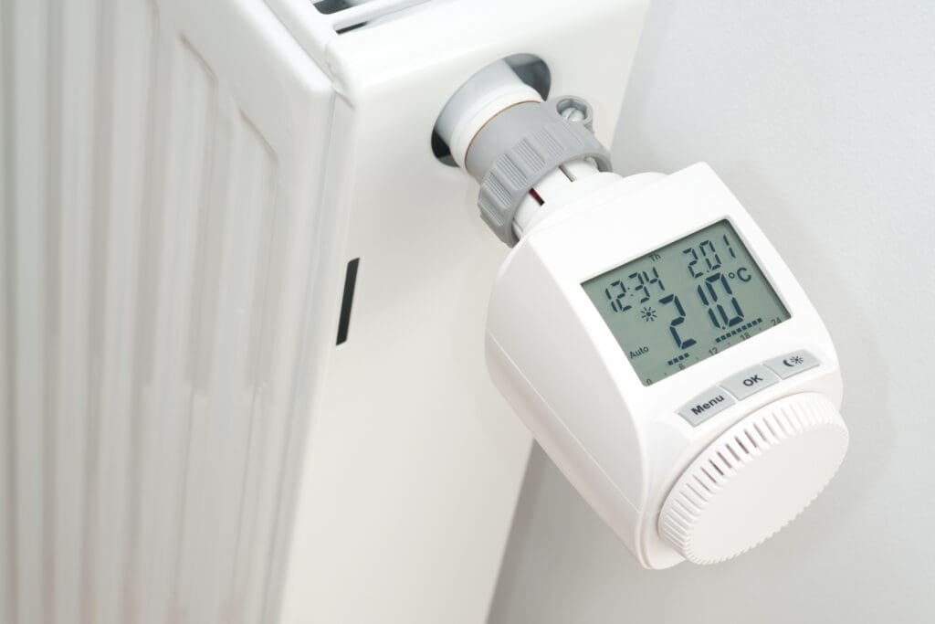 A close-up view of a digital thermostat attached to a white radiator, displaying the current temperature as 21.0°C, with buttons for menu and settings visible.