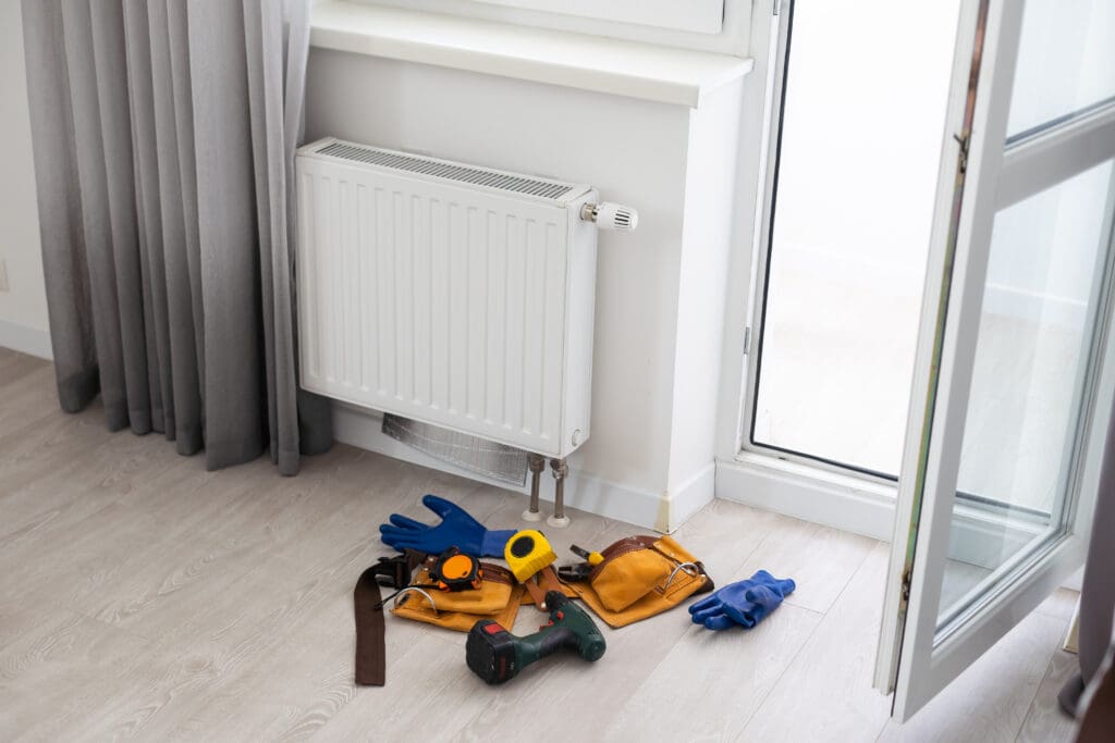 A set of tools, gloves, a measuring tape, and a tool belt are scattered on the floor near a white radiator under a window in a brightly lit room with an open door and curtains.