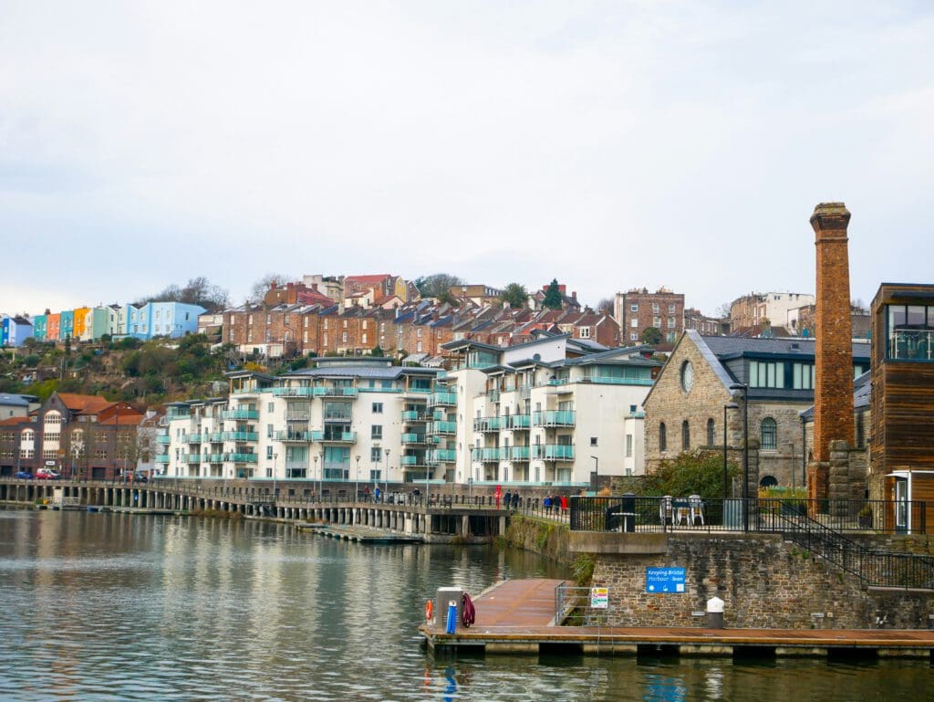 A waterfront scene with modern apartments, a historic brick building, and colorful houses on a hillside above the water under a cloudy sky.