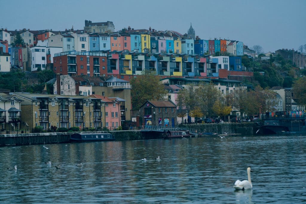 Colorful houses sit on a hillside above a river, with houseboats and birds in the water. A swan floats in the foreground, and trees with autumn leaves are near the riverbank. The sky is overcast.