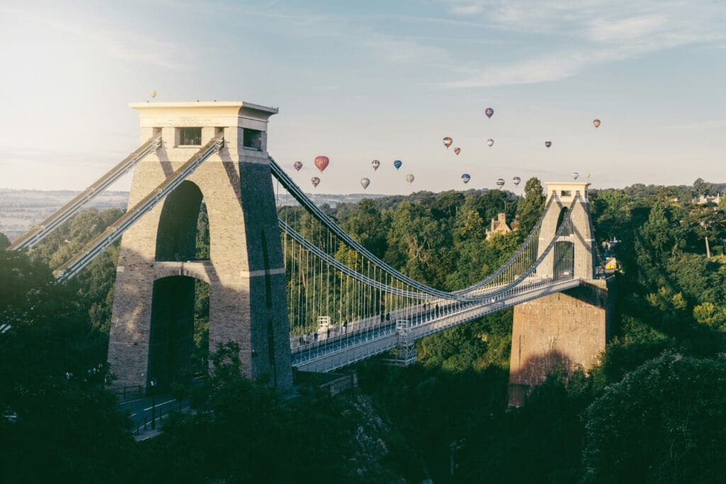 The Clifton Suspension Bridge spans a deep green gorge, with several colorful hot air balloons floating in the sky above a lush, tree-filled landscape on a clear day.