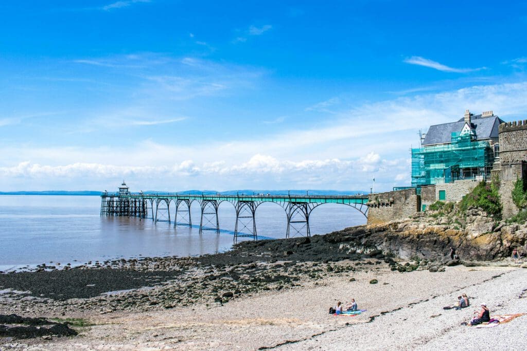 A Victorian pier extends over calm water from a rocky shoreline, with a few people relaxing on the pebbled beach. Nearby buildings, one under scaffolding, sit under a bright blue sky with scattered clouds.
