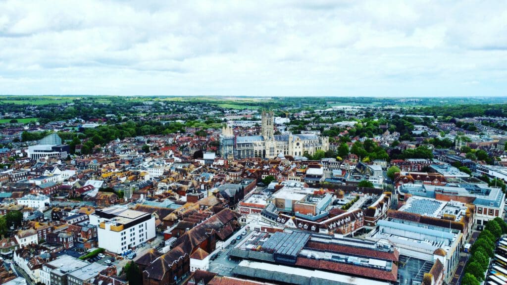 Aerial view of a historic city with dense, red-roofed buildings, a prominent cathedral at the center, and green fields in the distance under a cloudy sky.