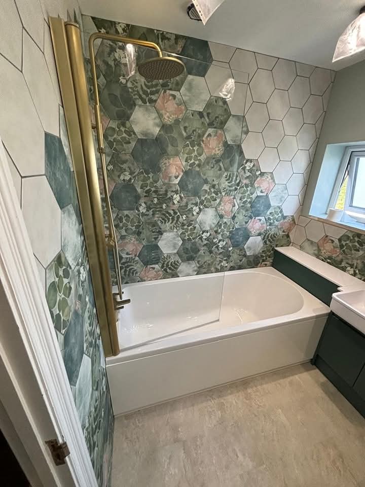 Modern bathroom with a white bathtub, green and floral hexagon wall tiles, a gold shower fixture, green vanity cabinets, and a small window letting in natural light.