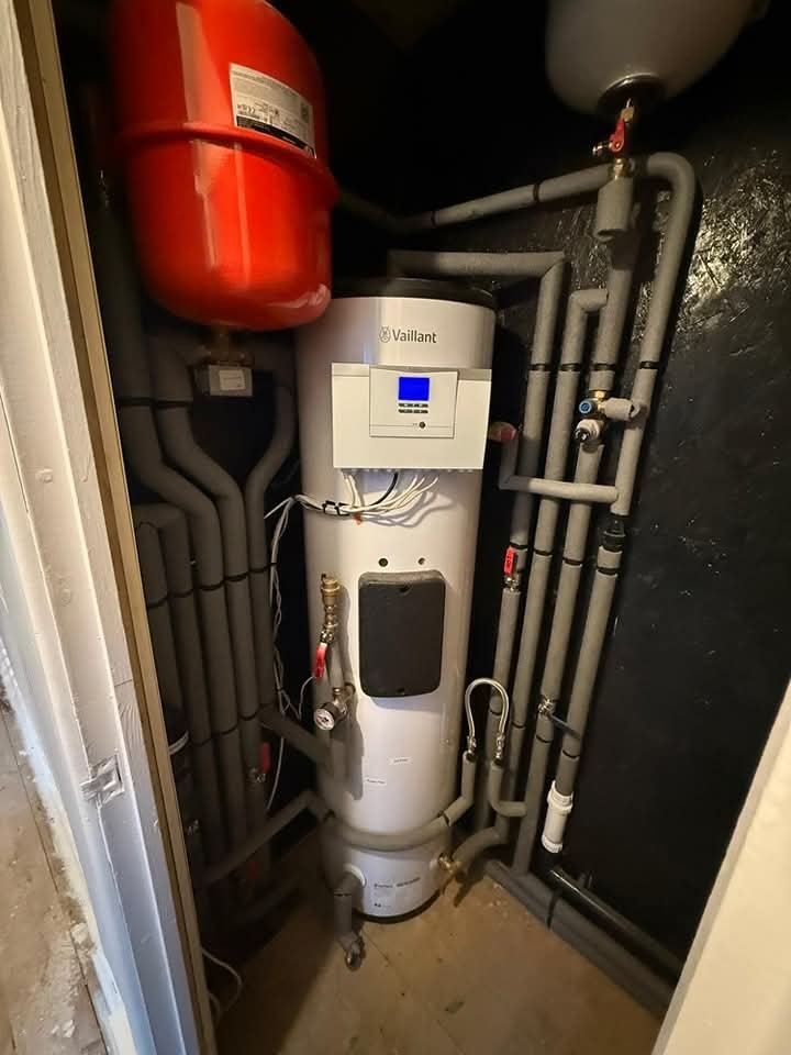 A modern hot water cylinder and heating system with a digital display is installed in a small utility closet, surrounded by insulated pipes and a large red expansion tank above.