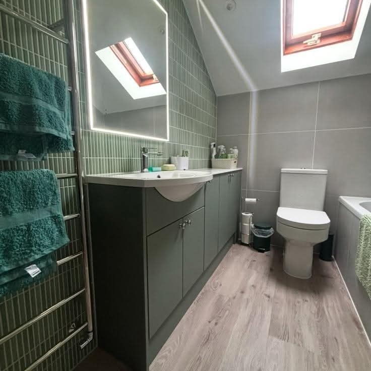 A modern bathroom with green tiled walls, a large mirror, grey cabinets, a white sink, a toilet, a towel rail with green towels, and a skylight above wooden flooring. Toiletries sit on the sink counter.