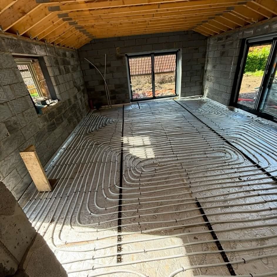 Room under construction with exposed walls and ceiling beams, featuring newly installed underfloor heating pipes laid out in loops across the insulated floor, with large windows letting in natural light.