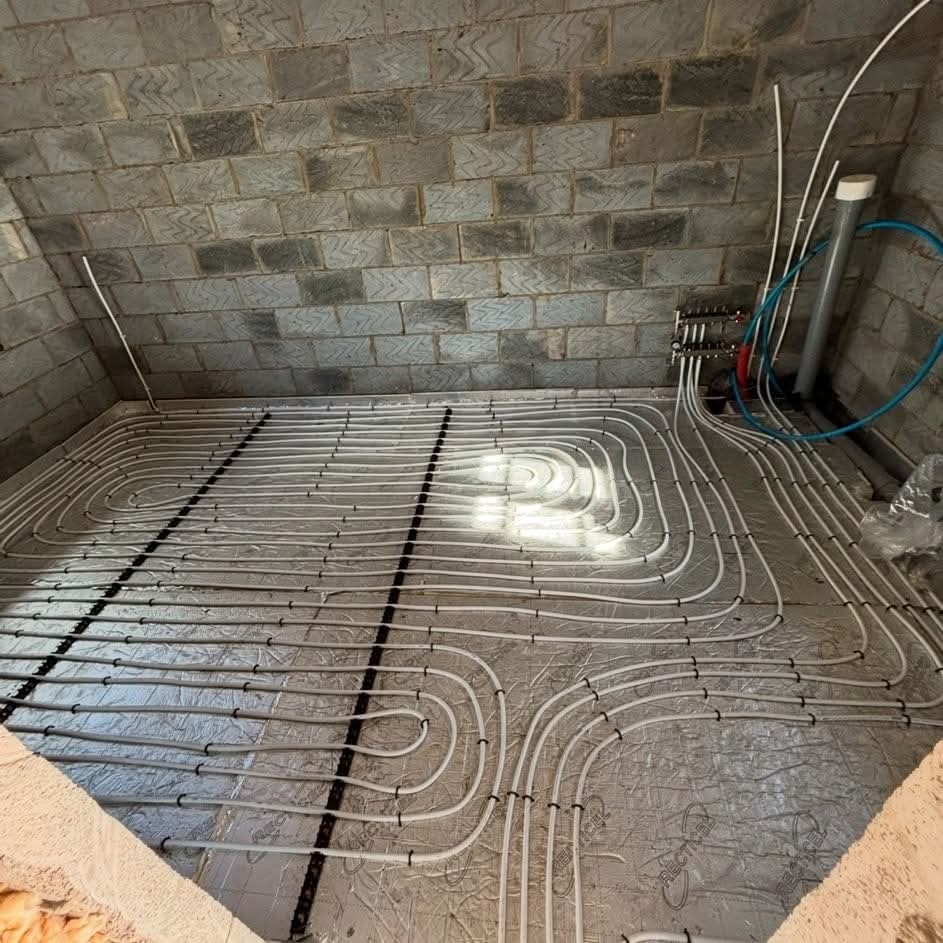 A room with unfinished concrete block walls showing an installed underfloor heating system, with white pipes arranged in loops across a foil-insulated floor and connected to a manifold in the corner.