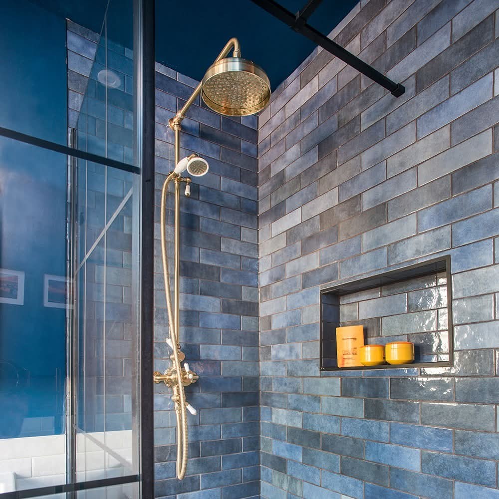 A modern shower with glossy blue subway tiles, a brass rain showerhead, and a built-in niche holding orange bottles and a yellow candle. A glass shower door is partially visible on the left.