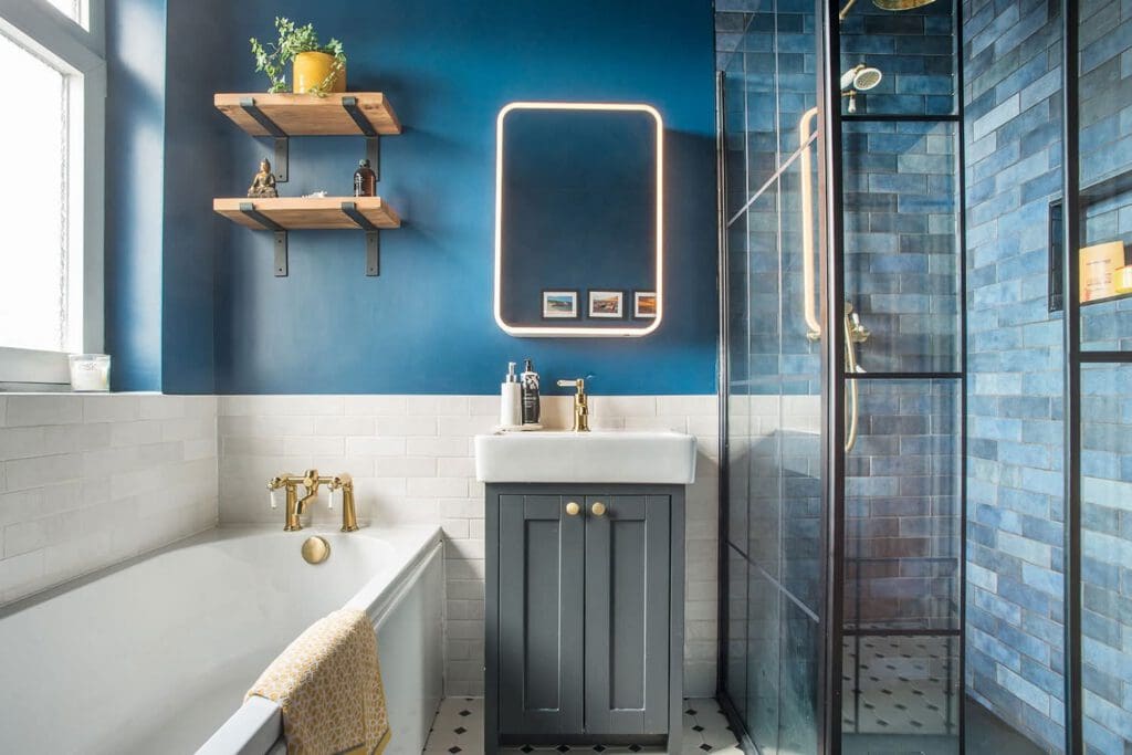 Modern bathroom with blue walls, a white bathtub with gold fixtures, a gray vanity, a lit mirror, wooden shelves with plants and bottles, and a glass shower with blue tiles and gold showerhead.