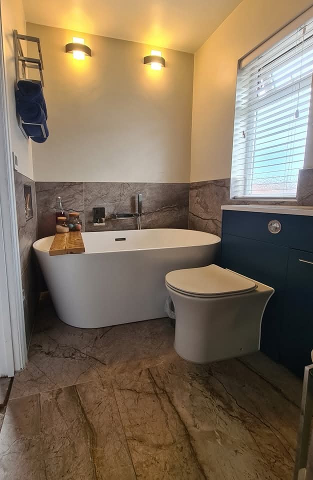 Modern bathroom with a white freestanding bathtub, a wooden bath tray, and a toilet next to a blue vanity unit under a window with blinds. The walls are partially tiled, and two wall lights are above the tub.