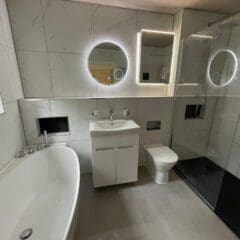 Modern bathroom with white marble walls, a bathtub, a sink with a cabinet, a toilet, and a walk-in shower. Two round, backlit mirrors are mounted above the sink and in the shower area.