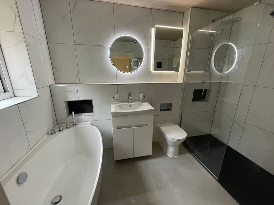 Modern bathroom with white marble tiles, a freestanding bathtub, a floating vanity with a round illuminated mirror, a toilet, and a walk-in shower with glass partition and another round backlit mirror.