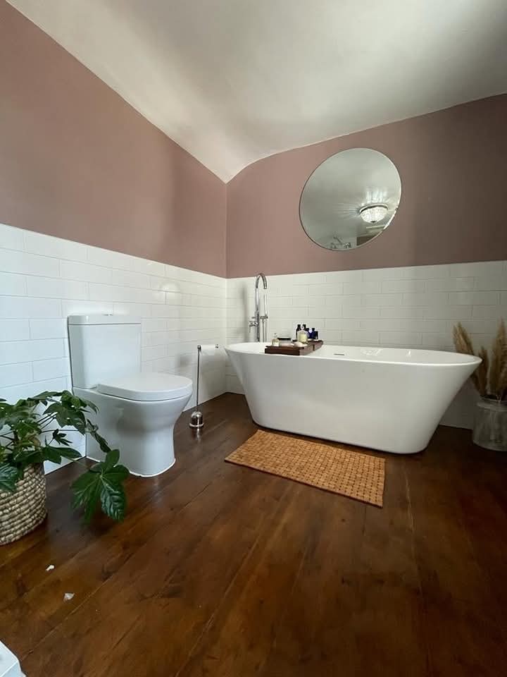 A modern bathroom with a white bathtub, toilet, and circular mirror. The walls are half white tile, half mauve paint, and the floor is wooden. A plant and a basket with pampas grass add natural accents.