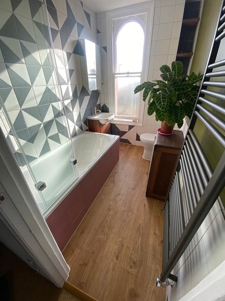 Modern bathroom with geometric tiled walls, a white bathtub with glass screen, wooden floor, toilet, small wooden cabinets, large leafy plant, and a tall arched window letting in natural light.