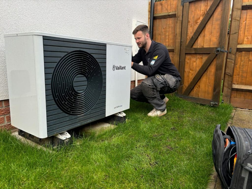 A technician kneels on grass while inspecting or installing a Vaillant outdoor heat pump unit near a house, with a wooden gate and a tool bag visible nearby.