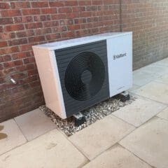 A Vaillant heat pump unit installed outside, positioned on a gravel bed next to a brick wall, with paved slabs surrounding the area.