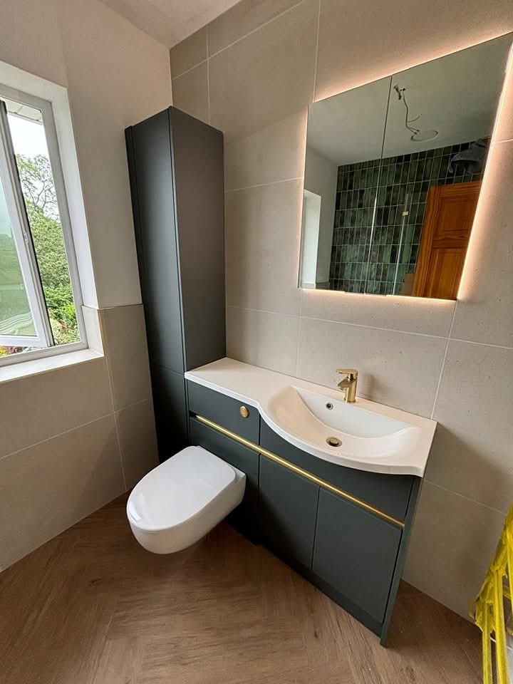 Modern bathroom with a wall-mounted toilet, dark vanity unit, white basin, gold faucet, and large illuminated mirror. Light tiles and wood-pattern flooring, with a window letting in natural light.