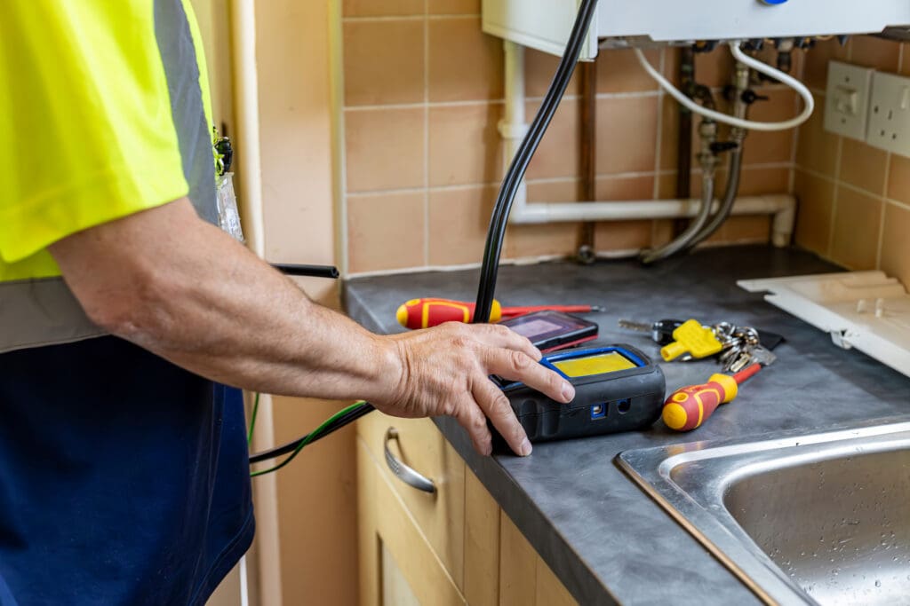A person tests a gas boiler in a kitchen using a digital analyzer on the countertop surrounded by tools, keys, and a phone, with pipes and a boiler unit visible in the background.