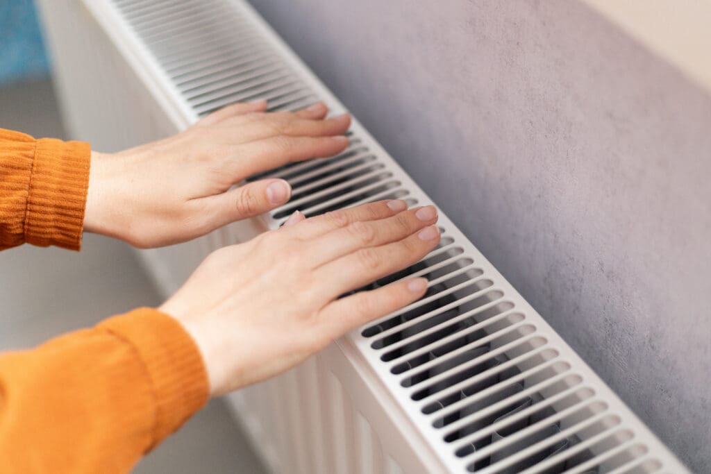 Two hands held above a white radiator, warming up. The person is wearing an orange long-sleeve top, and the radiator is against a light-colored wall.