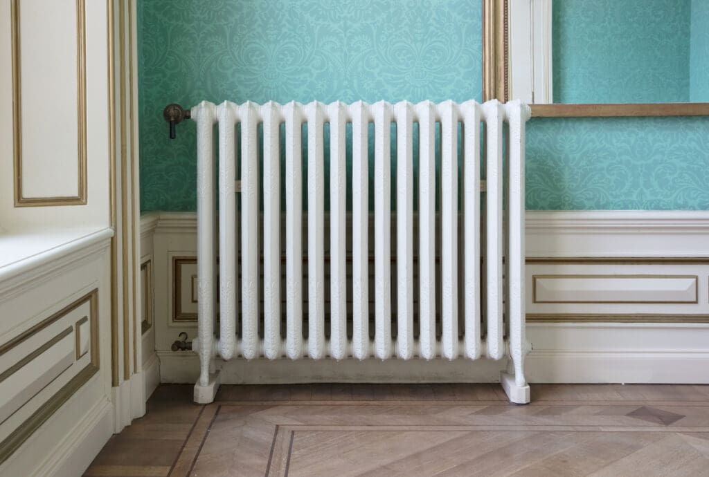 A white, vintage-style cast iron radiator stands against a teal wallpapered wall with ornate patterns in an elegant, classic room with white paneling and wood flooring.