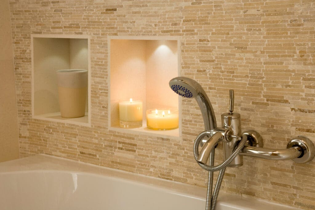 Close-up of a bathtub with a modern chrome faucet and handheld shower, set against a beige mosaic tile wall with built-in shelves holding candles and a ceramic cup, creating a relaxing atmosphere.