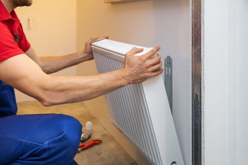 A person in blue pants and a red shirt installs a white radiator onto a wall bracket in a room. Tools, including a wrench and tape, are visible on the floor nearby.