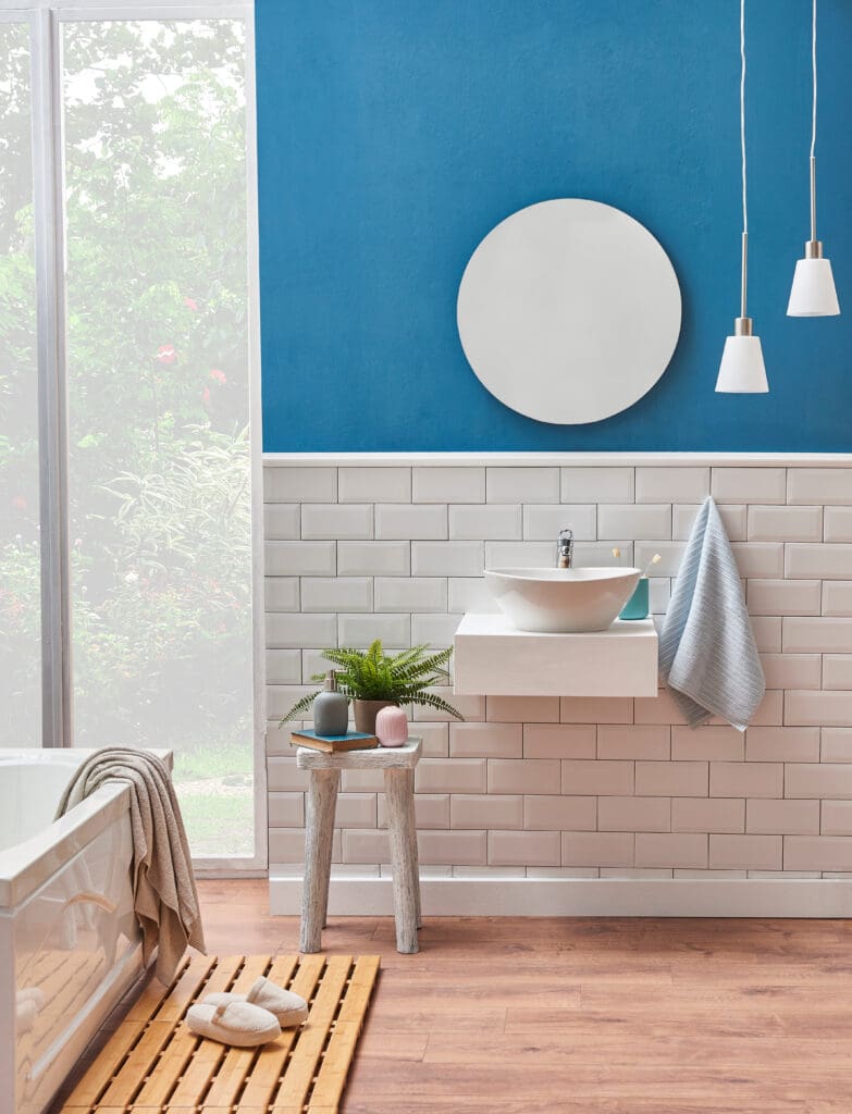 A modern bathroom with white subway tiles and a blue wall, featuring a round mirror, a floating sink, pendant lights, a stool with plants and decor, a towel, and a window showing greenery outside.