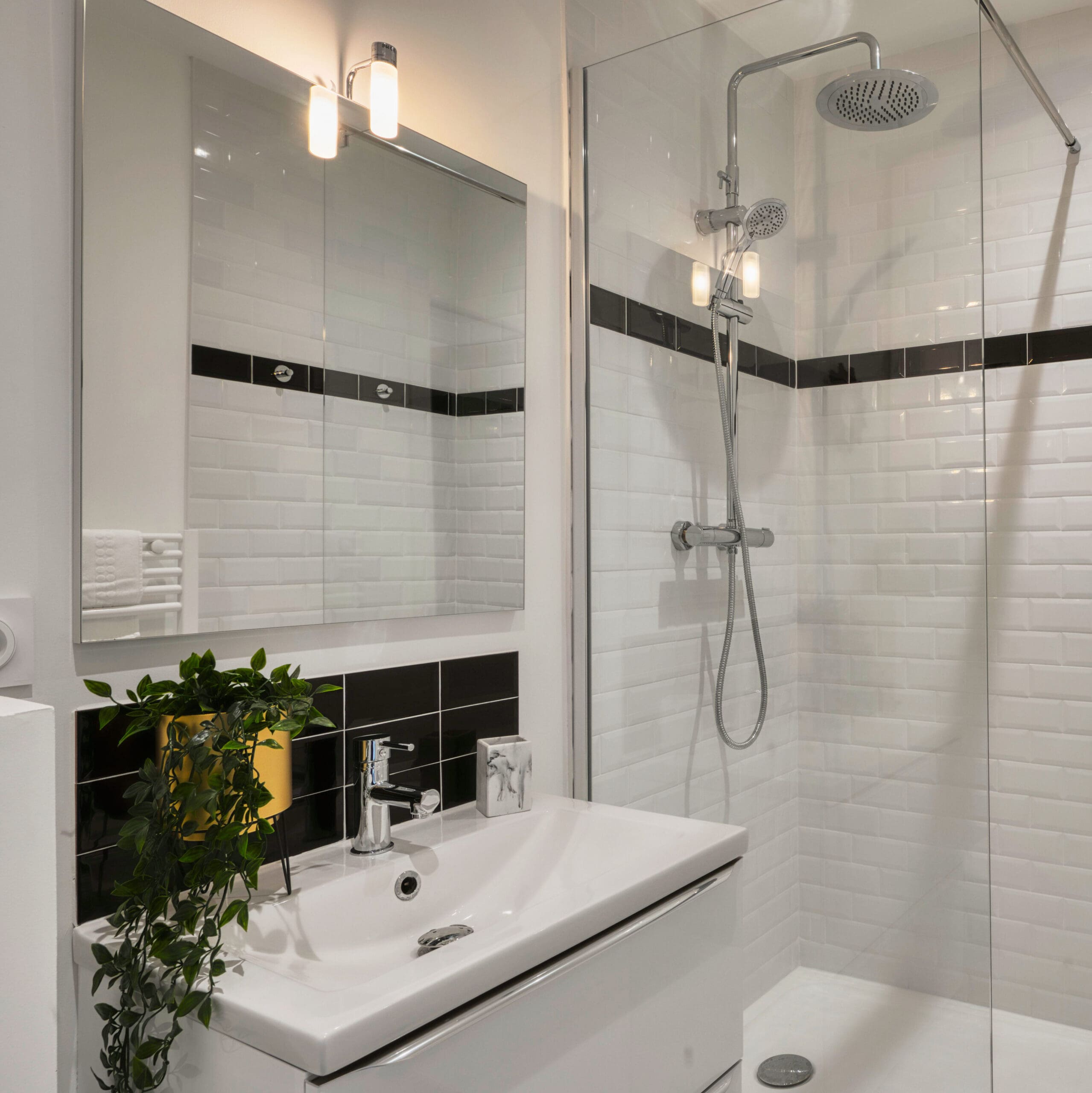 Modern bathroom with white subway tile walls, a glass shower enclosure, a rainfall showerhead, a rectangular sink with a chrome faucet, a mirror above the sink, and a green plant on the counter.